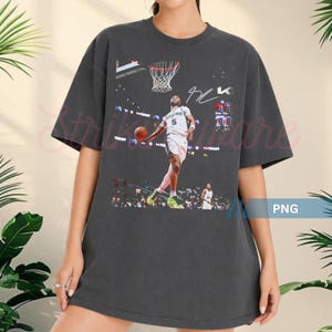 May include: Dark gray t-shirt featuring a basketball player in action, with a basketball and a hoop. The image includes a signature and the word "Tissot". The shirt is a casual, oversized fit.