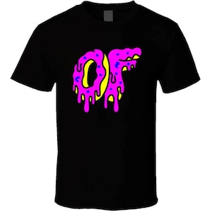 Può includere: T-shirt nero con una grafica delle lettere "OF" in un design a forma di ciambella. Le lettere sono rosa con centri gialli e spruzzi blu, con gocce rosa.
