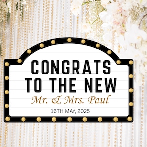 Può includere: Un cartello in bianco e nero con la scritta "CONGRATS TO THE NEW Mr. & Mrs. Paul 16TH MAY, 2025". Il cartello ha un bordo nero con accenti di lampadine dorate. Lo sfondo include fiori bianchi e fili di perline dorate.