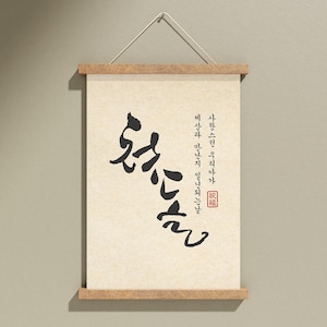 May include: A cream-colored scroll with Korean calligraphy, mounted on wooden bars and suspended by a white cord. The black ink script features vertical and horizontal text, with a red stamp at the bottom right. The wooden bars have a natural finish.