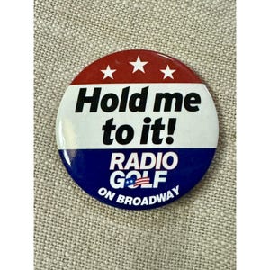 Puede incluir: Un botón redondo con un diseño rojo, blanco y azul. La sección superior es roja con tres estrellas blancas. La sección central es blanca con las palabras "Hold me to it!" en negro. La sección inferior es azul con el texto "RADIO GOLF ON BROADWAY".