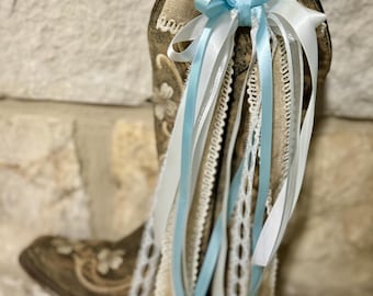 May include: A brown and tan cowboy boot decorated with white and light blue ribbons and lace. The ribbons are tied in a bow at the top of the boot. The boot has floral embroidery.