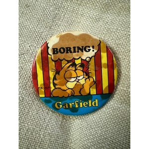 May include: A round Garfield pin with a cartoon image of Garfield looking bored. The pin has a red and yellow striped background, with the word "BORING!" in a thought bubble and the name "Garfield" at the bottom.