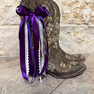 May include: Brown leather cowboy boots with floral embroidery. A purple satin bow and ribbons with white lace accents are attached to the boots. The boots are displayed on a tiled surface against a stone wall.