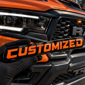 Puede incluir: Un emblema rectangular con fondo negro brillante y letras naranjas en relieve que forman la palabra CUSTOMIZED. El emblema se sostiene frente a la parrilla delantera naranja y el conjunto de faros de un camión RAM TRX.