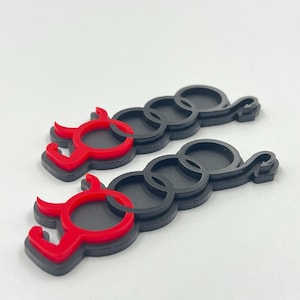 Puede incluir: Dos emblemas idénticos con cuatro anillos negros entrelazados, donde el anillo de la izquierda está modificado en rojo brillante para parecer una figura diabólica con cuernos y una cola puntiaguda. Diseño plano con bordes elevados.