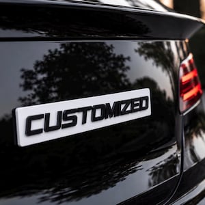 May include: A glossy black car trunk lid featuring a rectangular white emblem with the word CUSTOMIZED in bold, raised black lettering. The surface reflects the surrounding environment, and a portion of a red taillight is visible on the right side.