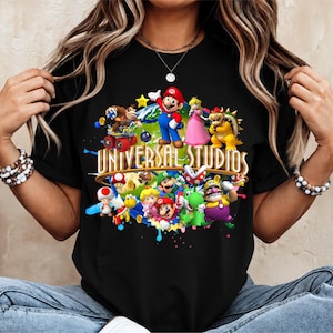 Könnte beinhalten: Ein schwarzes Kurzarm-T-Shirt mit einem farbenfrohen Grafikdruck verschiedener Nintendo-Figuren wie Mario, Luigi, Peach und Bowser, die einen Globus umgeben, mit dem Text UNIVERSAL STUDIOS in großen goldenen Buchstaben in der Mitte.