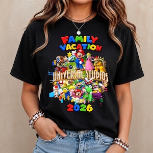 Könnte beinhalten: Ein schwarzes Kurzarm-T-Shirt mit einem bunten Grafikdruck und dem Text FAMILY VACATION UNIVERSAL STUDIOS 2026. Das Design zeigt verschiedene Videospielcharaktere wie Mario, Luigi, Prinzessin Peach, Bowser und Donkey Kong.