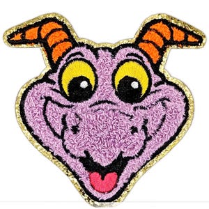 Könnte beinhalten: Gestickter Aufnäher mit dem Gesicht einer lila Kreatur mit gelben Augen, orangefarbenen Hörnern und einer rosa Zunge. Der Aufnäher hat einen goldenen Glitzerrahmen und eine flauschige Textur. Das Design ist im Cartoon-Stil.