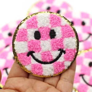 Op de afbeelding: Een ronde, geborduurde patch met een smiley-gezichtontwerp. Het gezicht heeft een roze en wit geblokt patroon, zwarte ogen en een zwarte glimlach. De patch is omrand met gouden pailletten.
