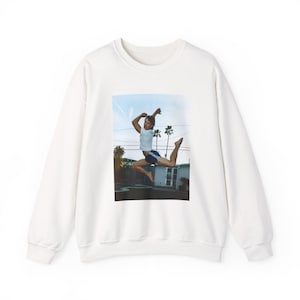 Peut inclure: Sweat-shirt blanc à col rond avec une image imprimée d'une personne en l'air, portant un t-shirt blanc et un short bleu. L'image a une toile de fond de palmiers et une maison sous un ciel bleu. Vêtement décontracté sur le thème du sport.