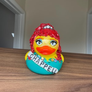 May include: A novelty rubber duck with a yellow body, orange beak, and teal dress. The duck features red curly hair, a silver tiara, and a sash reading "CHAPPELL". Rhinestones decorate the sash and dress. A quirky and fun collectable item.