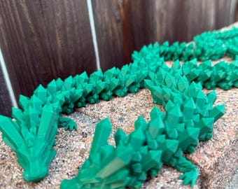 May include: A vibrant green, 3D-printed dragon figurine with a crystalline design. The dragon is segmented, with a head, body, and tail. The figurine is resting on a textured, light brown surface, with a wooden fence in the background.