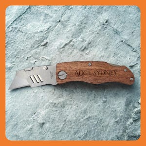 May include: A utility knife with a wooden handle and a stainless steel blade. The handle is engraved with the text "ALICE SYDNEY". The blade is silver and has three rectangular cutouts. The knife is approximately 6 inches long.