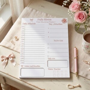 May include: A Daily Bloom time-blocking planner with a daily schedule, top priorities, to-do list, self-care, notes, and water tracker. A pink pen, floral mug, and roses are on a table.