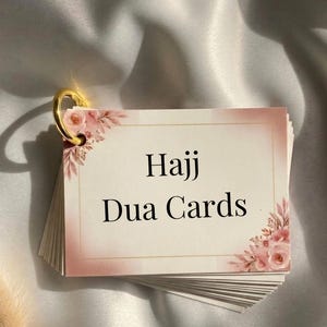 Peut inclure: Un ensemble de cartes Hajj Dua, maintenues par un anneau doré. Les cartes présentent un motif floral avec des roses roses et les mots "Hajj Dua Cards". Le fond est de couleur neutre.
