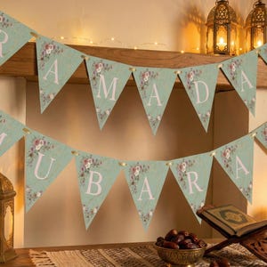May include: A festive banner with the words "RAMADAN MUBARAK" in white letters on mint green pennants, adorned with floral patterns. The scene includes lit lanterns, candles, and a bowl of dates, creating a warm, celebratory atmosphere.