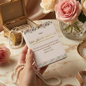 Può includere: Una carta decorativa con accenti floreali e testo arabo è tenuta vicino a una collana di perle e a uno scrigno d'oro. La carta recita "Giorno 3" e include una citazione del Corano. Una tazza di tè e rose sono sullo sfondo.