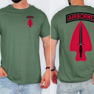 May include: Olive green t-shirt featuring a red and black emblem with a dagger and the word "AIRBORNE". The emblem is on the front and back of the shirt. The shirt is a crew neck and has short sleeves.