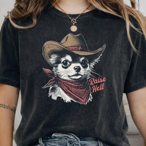 May include: A charcoal black short-sleeve t-shirt featuring a graphic of a chihuahua wearing a brown cowboy hat and a red patterned bandana. The text "Raise Hell" is printed in red script next to the dog. The shirt has a vintage washed finish.