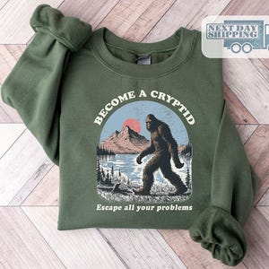 May include: An olive green crewneck sweatshirt featuring a graphic print of a Bigfoot creature walking near a lake and mountains. The text above the graphic reads BECOME A CRYPTID and the text below reads Escape all your problems.