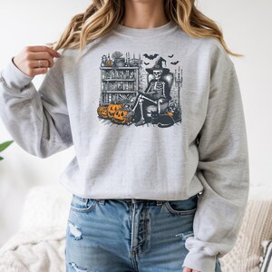 May include: A light gray crewneck sweatshirt featuring a graphic print of a skeleton wearing a witch hat, sitting in a chair reading a book. The illustration includes a bookshelf, orange pumpkins, a black cat, and bats in a black and orange ink style.