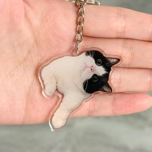 Op de afbeelding: Een heldere acryl sleutelhanger met een zwart-wit kattenontwerp. De kat ligt met zijn hoofd gekanteld, met zwarte oren en ogen en een roze neus. De sleutelhanger heeft een zilveren ketting en ring.