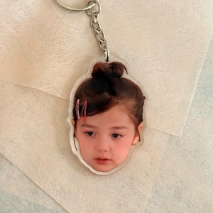 May include: A clear acrylic keychain featuring a young girl's face. The keychain has a silver metal ring and chain. The girl has brown hair styled in a bun with red barrettes. The background is a light, textured paper.