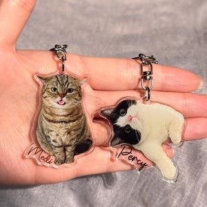 May include: Two cat-shaped keychains. One keychain features a tabby cat with the name "Medi" printed on it. The other keychain features a black and white cat with the name "Percy" printed on it. Both keychains have silver-colored key rings.