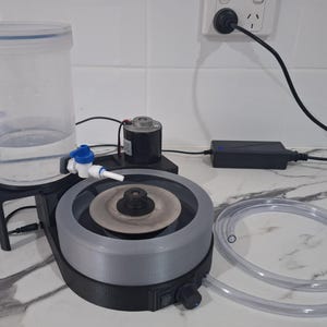 May include: A lapidary machine featuring a circular grey grinding disc, a black base with a power switch and speed control knob, a clear plastic water reservoir with a blue valve, a small motor, a power adapter, and a clear flexible tubing.