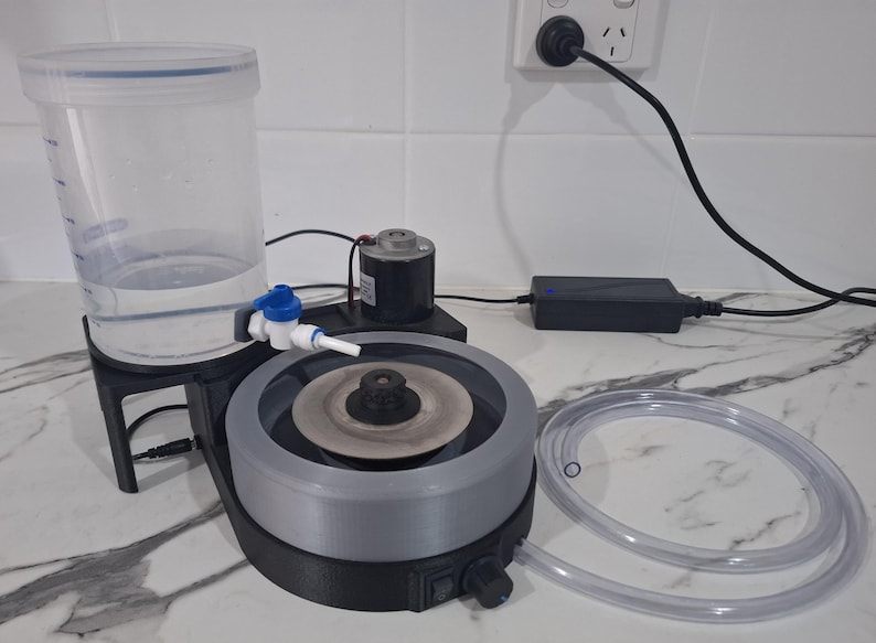 May include: A lapidary machine featuring a circular grey grinding disc, a black base with a power switch and speed control knob, a clear plastic water reservoir with a blue valve, a small motor, a power adapter, and a clear flexible tubing.