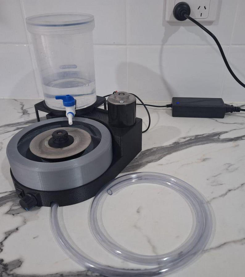 May include: A black and grey lapidary machine featuring a circular grinding disc, a clear plastic water reservoir with a blue valve, a motor, and a long, flexible, transparent plastic hose. The device includes a black power adapter with a cord.