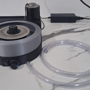 May include: A black and grey electric lapidary machine featuring a circular grinding disc, a motor, a power switch, and a control knob. A clear plastic hose is attached to the side of the base, and a black power adapter is connected by a cord.