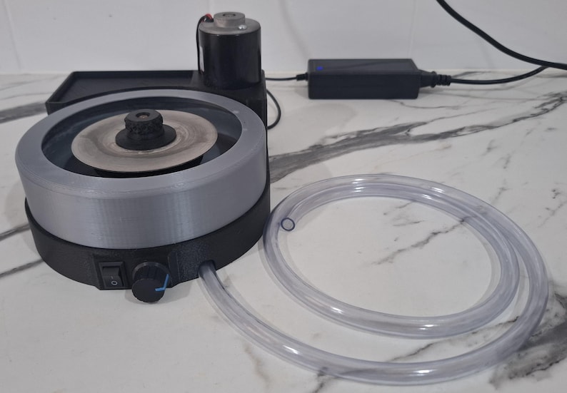 May include: A black and grey electric lapidary machine featuring a circular grinding disc, a motor, a power switch, and a control knob. A clear plastic hose is attached to the side of the base, and a black power adapter is connected by a cord.