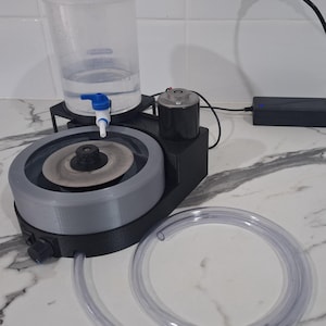 May include: A black and grey lapidary machine featuring a circular grinding disc, a clear plastic water reservoir with a blue spigot, a black motor housing, and a long, flexible clear plastic drainage tube connected to the base unit.