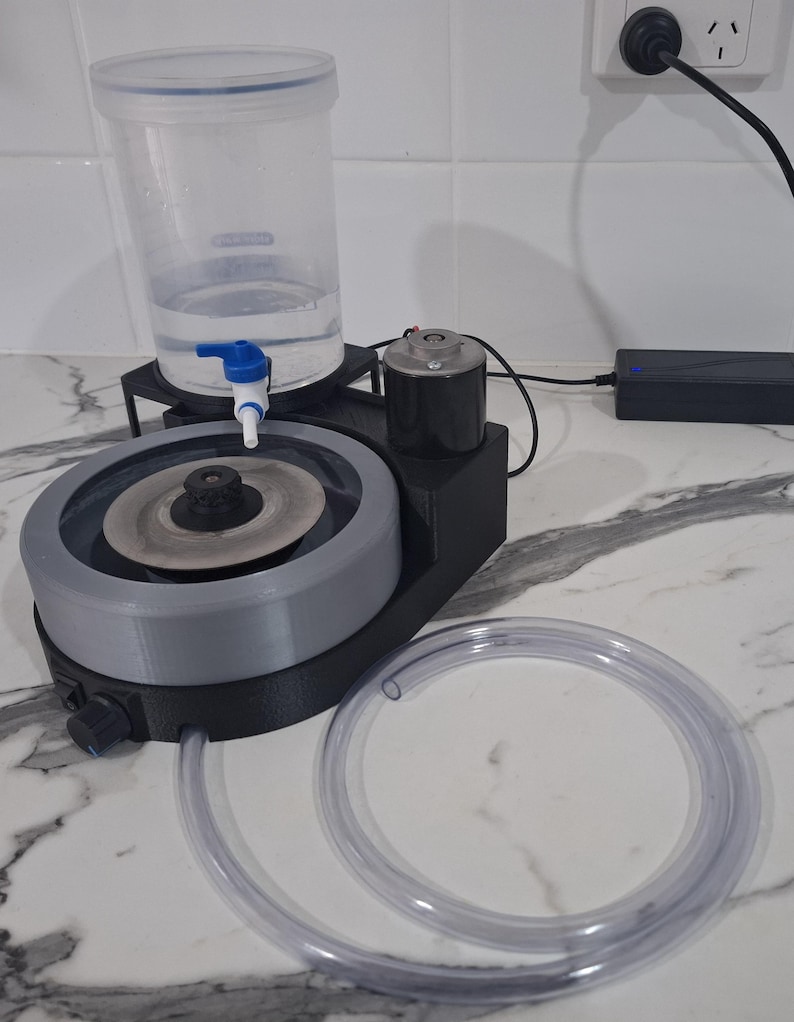 May include: A black and grey lapidary machine featuring a circular grinding disc, a clear plastic water reservoir with a blue spigot, a black motor housing, and a long, flexible clear plastic drainage tube connected to the base unit.