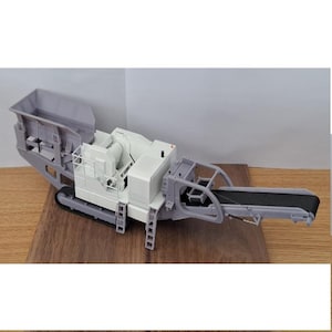 May include: A model of a heavy-duty industrial machine, primarily in shades of gray and white, with a black conveyor belt. The machine has a hopper, a central processing unit, and a conveyor belt for material transport. The model is set on a wooden surface.