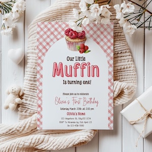 May include: A birthday invitation with a pink gingham pattern and a cupcake illustration topped with raspberries. The text reads "Our Little Muffin Is turning one!" and includes party details. The invitation is surrounded by white flowers and a soft blanket.