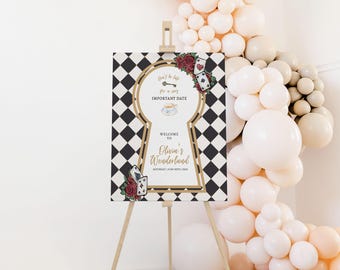 May include: A decorative sign on a wooden easel with a black and white checkered background. The sign features a keyhole design with the text "Welcome to Olivia's Wonderland" and floral and playing card accents. A balloon arch is in the background.