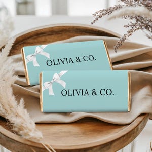 May include: Two light blue rectangular bars with white bow detail and the text "OLIVIA & CO." in black. The bars have gold edges and are presented on a wooden tray with a neutral-colored cloth.