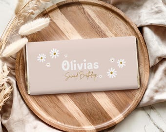 Op de afbeelding: Een lichtroze chocoladereep met de tekst "Olivias Second Birthday" in wit en goud. De reep is versierd met witte madeliefjes en wordt gepresenteerd op een houten dienblad.