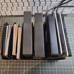 May include: A collection of black and gray electronic devices, including what appears to be a card reader, a stack of cards, and several rectangular boxes. The items are arranged on a yellow grid surface, with a keyboard in the background.
