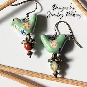 May include: A pair of handmade earrings featuring small, light green bird-shaped ceramic charms with orange and blue accents. Each earring has a red bead and a smaller, dark bead hanging below. The text "Designs by Jewelry Petaling" is visible.