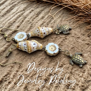 Puede incluir: Pendientes colgantes con cuentas de concha, detalles de flores blancas y dijes de tortugas marinas de bronce. Los pendientes tienen ganchos dorados y se exhiben sobre una superficie arenosa. El texto "Designs by Jewelry Petaling" es visible.