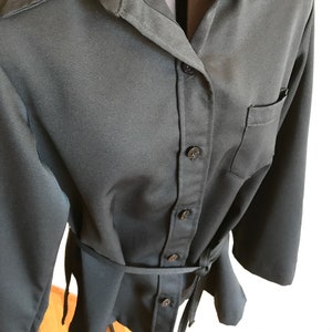 May include: A black long-sleeved shirt with a button-up front and a tie belt. The shirt has a collar and a pocket on the left side.