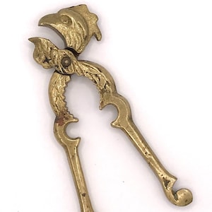 May include: A gold-toned brass nutcracker shaped like a bird with intricate details. The nutcracker has a hinged jaw and a lever to crack nuts.