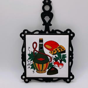 May include: Black cast iron trivet with a decorative tile featuring a wine bottle, cheese, and radishes. The tile has a white background with colorful illustrations. The trivet has a handle for easy use.