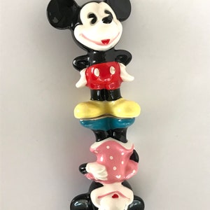 May include: A ceramic figurine of Mickey Mouse and Minnie Mouse stacked on top of each other. Mickey is wearing red shorts and yellow shoes. Minnie is wearing a pink polka dot dress and yellow shoes.