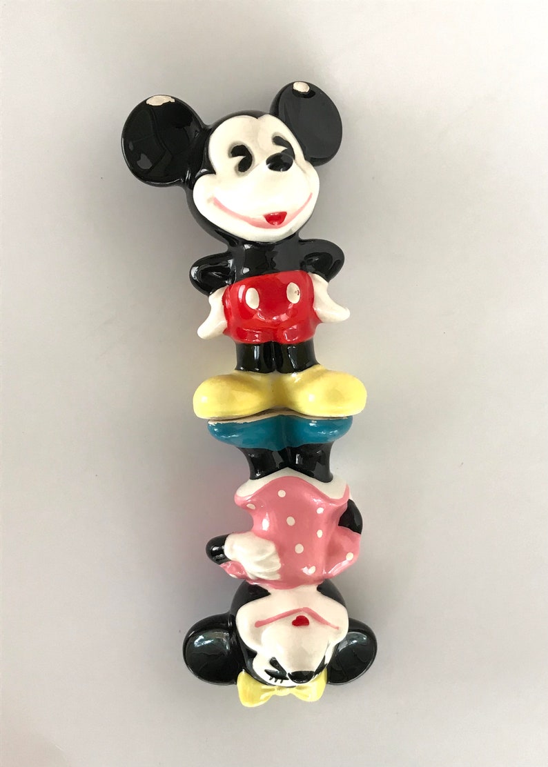 May include: A ceramic figurine of Mickey Mouse and Minnie Mouse stacked on top of each other. Mickey is wearing red shorts and yellow shoes. Minnie is wearing a pink polka dot dress and yellow shoes.